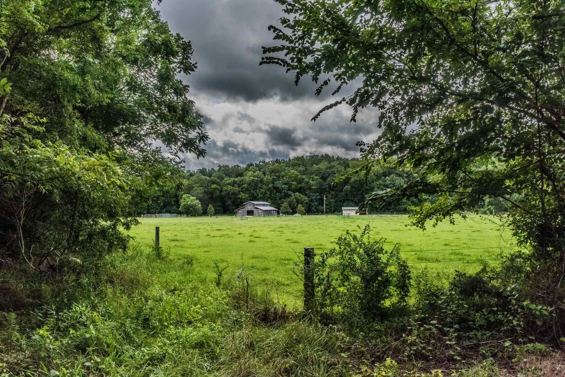 Green pasture with a barn in the distance and wooded hills behind under dramatic storm clouds